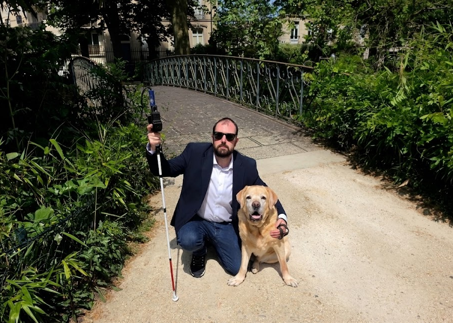 Kévin Derome, accroupi sur un petit pont dans un parc ensoleillé, tient sa canne blanche équipée d'ICane ; son chien-guide golden retriever est assis à côté de lui.